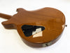 Back view of a polished wooden electric guitar body with a black plastic control cavity cover and metal output jack plate on white background