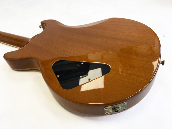 Back view of a polished wooden electric guitar body with a black plastic control cavity cover and metal output jack plate on white background