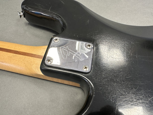Close-up of black electric guitar body showing worn finish, engraved metal neck plate with four screws, and maple neck with walnut stripe on gray background.