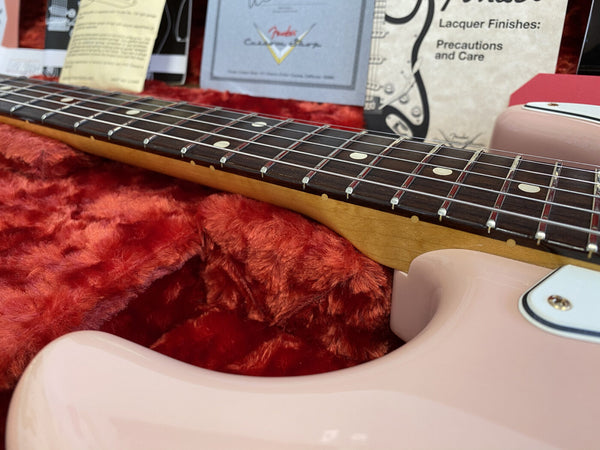 Close-up of pink electric guitar neck and fretboard with dot inlays resting on red plush case lining with Fender care pamphlets in background