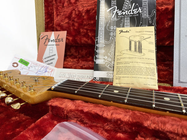 Close-up of Fender electric guitar neck and headstock with gold tuning pegs, inside red plush guitar case, showing Fender instruction manuals and documentation for tremolo springs.