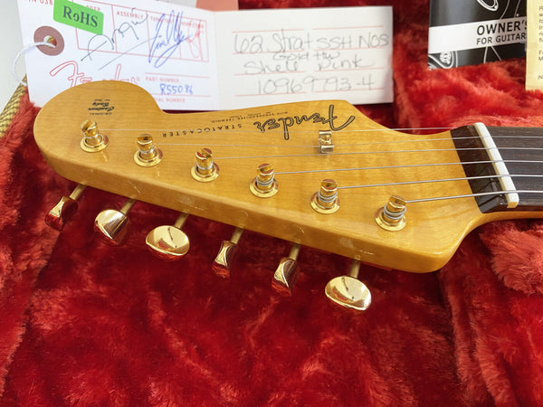 Close-up of Fender Stratocaster guitar headstock with gold tuning pegs and strings on red plush background, vintage 62 Strat SSH NOS model with original Fender tag and owner's manual visible