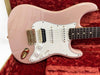 Pink electric guitar with white pickguard, gold hardware, and rosewood fingerboard on red plush guitar case interior.