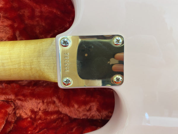 Close-up of guitar neck plate with serial number R55086 on light-colored wood neck and white guitar body, resting on red plush guitar case lining