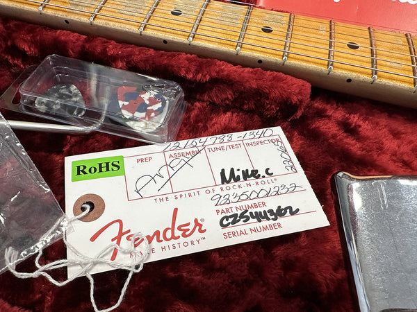 Close-up of Fender guitar neck with maple fretboard, Fender branded inspection tag with part and serial numbers, guitar picks in plastic case, and chrome guitar bridge plate on red plush guitar case lining.