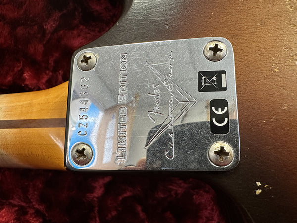 Close-up of Fender Custom Shop limited edition serial number C7544362 metal neck plate on electric guitar with CE certification and no battery disposal symbols on dark brown guitar body, photographed on red textured surface