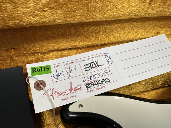 Close-up of Fender guitar inspection tag with RoHS label, serial number R74690, and handwritten prep, assembly, and inspector details, resting on gold fabric lining and part of black and white guitar body visible.
