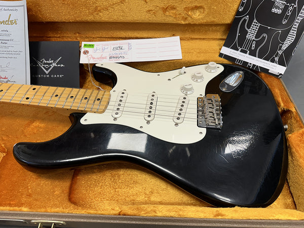 Black Fender Stratocaster electric guitar with maple neck and white pickguard, displayed in open orange plush guitar case with Fender Custom Care documents and certification cards