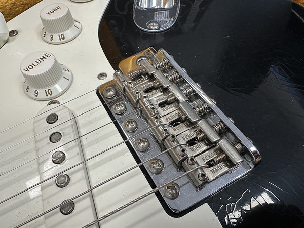 Close-up of black Fender electric guitar bridge, white volume and tone control knobs, and strings on white pickguard with metal saddles marked Fender.