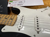 Close-up of Fender Stratocaster electric guitar white pickguard, two single-coil pickups, maple fretboard, and part of Fender quality control tag with serial number and inspector signatures.