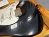 Close-up of black electric guitar body with white pickguard, volume and tone knobs, and metal bridge, showing wear and cracks in finish inside a plush orange guitar case