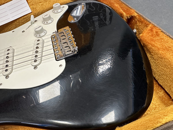 Close-up of black electric guitar body with white pickguard, volume and tone knobs, and metal bridge, showing wear and cracks in finish inside a plush orange guitar case