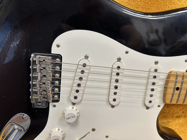 Close-up of black electric guitar body focusing on white pickguard, three single-coil pickups, volume and tone knobs, and chrome bridge with six strings