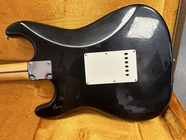 Black electric guitar body with visible wear and finish cracks, white backplate, and metal neck plate, placed in orange plush guitar case