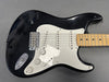Black electric guitar with white pickguard, three single-coil pickups, maple fretboard, and volume and tone control knobs on gray background