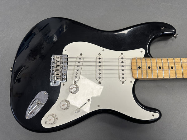 Black electric guitar with white pickguard, three single-coil pickups, maple fretboard, and volume and tone control knobs on gray background