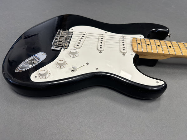 Black electric guitar with white pickguard and maple neck shown on gray surface, close-up view of volume and tone knobs, pickup switches, and bridge hardware.