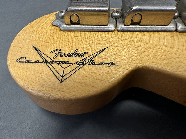 Close-up of Fender Custom Shop logo on natural wood guitar headstock with metal tuning machines