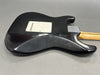 Black electric guitar body with white backplate and natural wood neck on gray surface, showing textured and worn finish