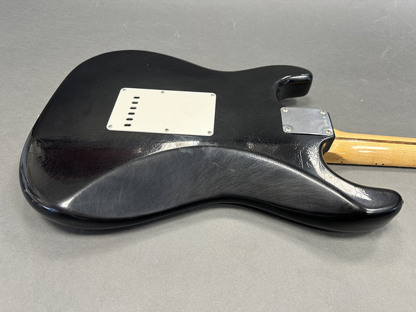 Black electric guitar body with white backplate and natural wood neck on gray surface, showing textured and worn finish