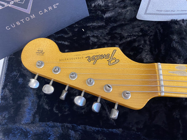 Close-up of Fender Stratocaster guitar headstock with six tuning pegs, maple wood finish, and Fender logo, resting on black plush guitar case interior with custom care booklet visible.