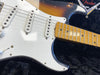 Close-up of sunburst electric guitar body, white pickguard, three single-coil pickups, volume control knob, and maple fretboard with black dot inlays on black plush case with Fender logo.
