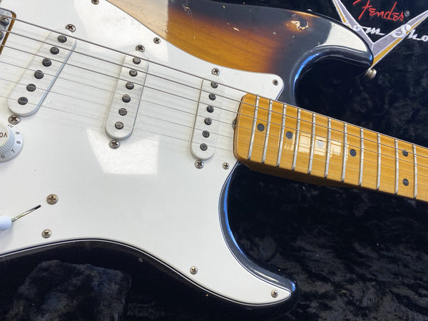 Close-up of sunburst electric guitar body, white pickguard, three single-coil pickups, volume control knob, and maple fretboard with black dot inlays on black plush case with Fender logo.