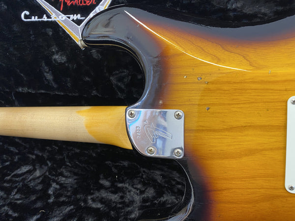 Close-up of Fender Custom electric guitar neck joint showing sunburst finish, chrome neck plate with Fender logo, and aged wear marks on guitar body and maple neck against black plush case interior.