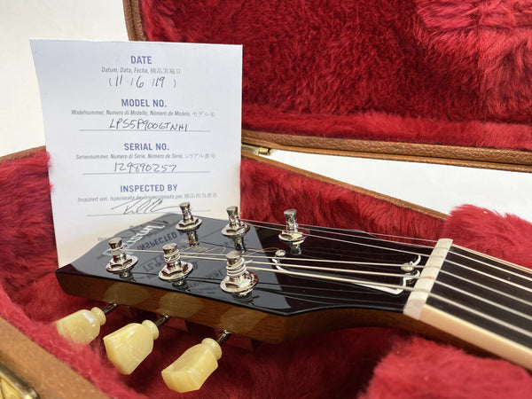 Close-up of Gibson Les Paul guitar headstock with tuning pegs and inspection certificate inside red plush-lined guitar case