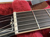 Close-up image of guitar nut, fretboard, and strings resting on red plush case lining with a leather Gibson strap in the background