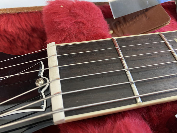 Close-up image of guitar nut, fretboard, and strings resting on red plush case lining with a leather Gibson strap in the background