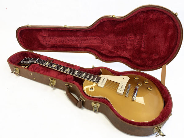 Gold top Les Paul electric guitar in open brown hard case with red plush interior lining, showing guitar body and neck detail with cream pickguard and knobs