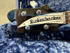 Close-up of black Rickenbacker bass guitar headstock with silver tuning pegs and "Rickenbacker Made in USA" logo on blue textured fabric background