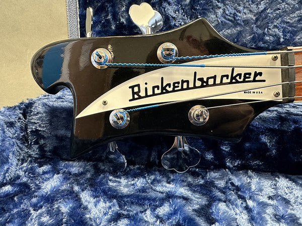 Close-up of black Rickenbacker bass guitar headstock with silver tuning pegs and "Rickenbacker Made in USA" logo on blue textured fabric background