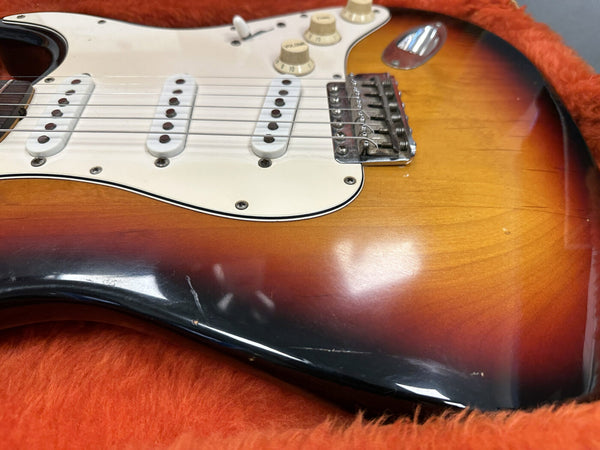 Close-up of sunburst electric guitar body with three single-coil pickups, white pickguard, and volume and tone knobs, resting on an orange fuzzy surface