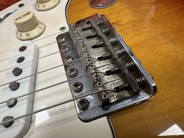 Close-up of vintage Fender electric guitar bridge, saddles, and strings on sunburst body with volume knobs and white pickguard