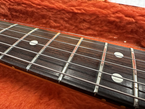Close-up of guitar fretboard with metal frets and white dot inlays on a dark wood fingerboard against an orange textured background