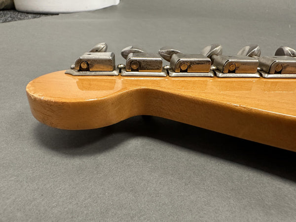 Close-up of vintage guitar headstock with six metal tuning machines, natural wood finish, and aged hardware, shown on gray background.