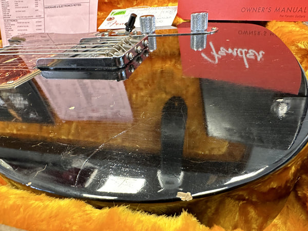 Close-up of black electric guitar body showing scratches and wear, with part of the bridge and control knobs visible, resting on a yellow plush case interior with Fender owner's manual in the background.