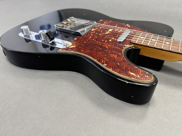 Close-up angled view of black electric guitar with tortoiseshell pickguard and chrome control knobs on gray background