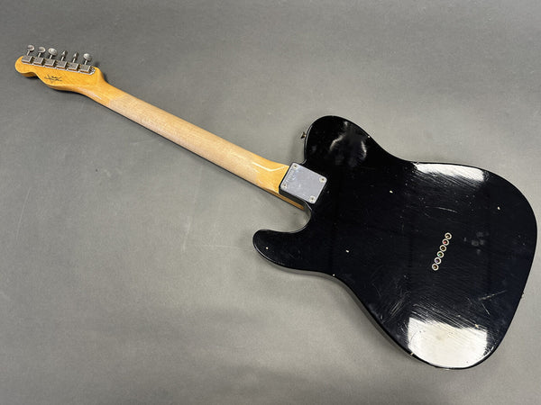 Back view of black electric guitar with maple neck and vintage-style tuners on gray background, showing string ferrules and metal neck plate.