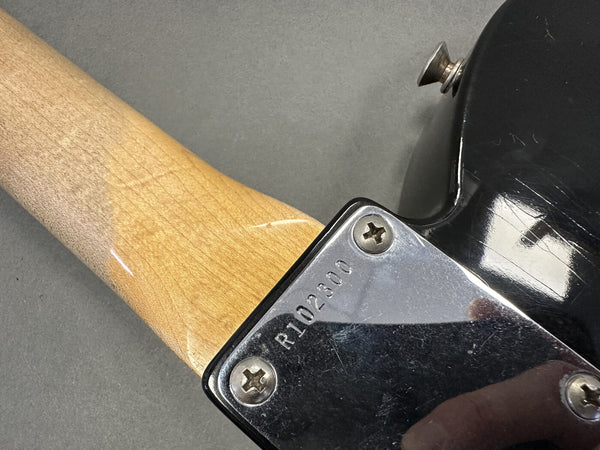 Close-up of guitar neck joint showing serial number R1102300 on metal plate with screws on a black electric guitar body and natural wood neck.