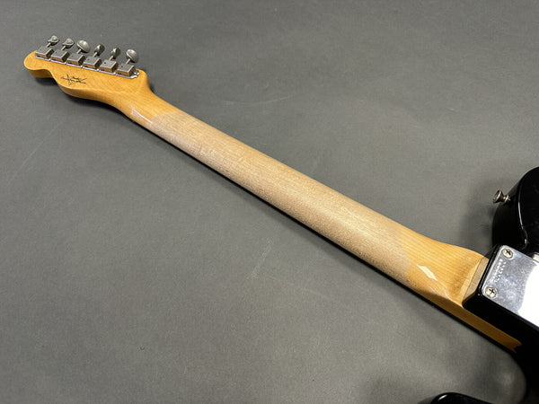Back side of a Fender electric guitar neck showing tuning pegs, maple wood neck, and black guitar body on gray background.