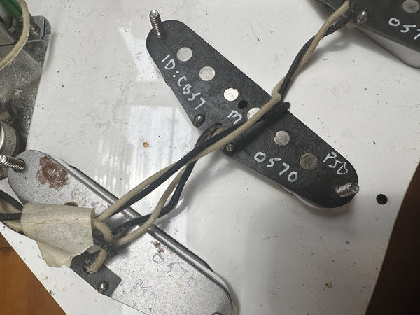 Close-up of vintage electric guitar pickups with handwritten identification numbers and cloth-covered wiring on a white surface at Coffee House Guitars