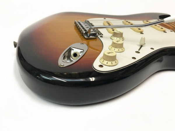 Close-up of sunburst electric guitar body showing chrome output jack plate, white tone and volume control knobs, and whammy bar on white pickguard against white background