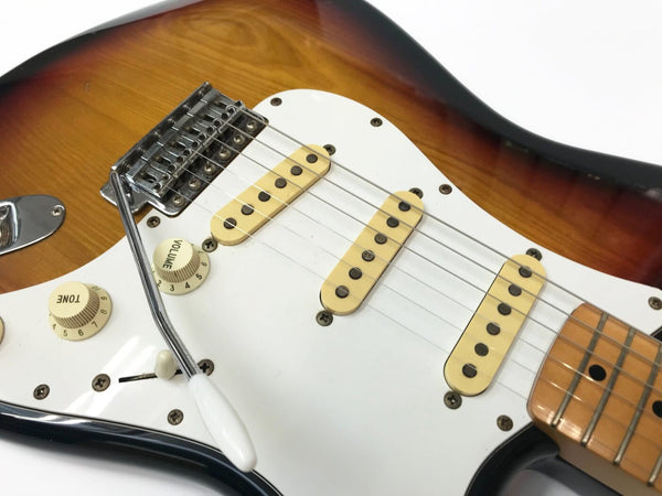 Close-up of sunburst electric guitar body showing white pickguard, three single-coil pickups, volume and tone knobs, and tremolo arm with maple neck fretboard