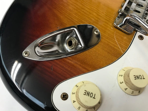Close-up of sunburst electric guitar body showing chrome output jack plate, white tone control knobs, and white pickguard with scratches and wear.