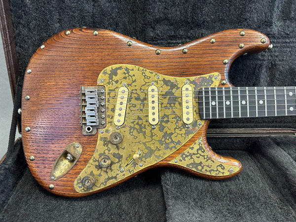 Close-up of custom electric guitar with natural wood finish and vintage brass pickguard featuring three single-coil pickups and metal hardware screws, displayed in a black padded guitar case