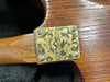 Close-up of a brass neck plate on a handmade Pajéttie Guitars electric guitar with visible wood grain and neck serial number engraved on the neck heel.