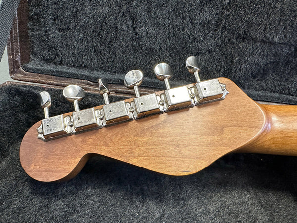 Back view of guitar headstock with six chrome tuning machines, vintage style, on natural wood neck resting in black plush guitar case